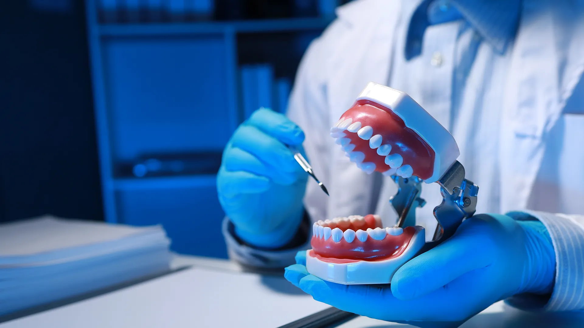 Dental professional in blue gloves adjusting a set of complete dentures on a model, illuminated under blue lighting to emphasize precision and detail in denture fitting.