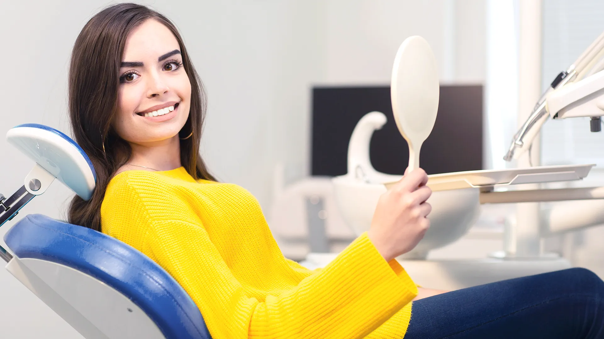 Young woman in a bright yellow sweater sitting in a dental chair, smiling as she examines her teeth in a handheld mirror, in a modern dental office.