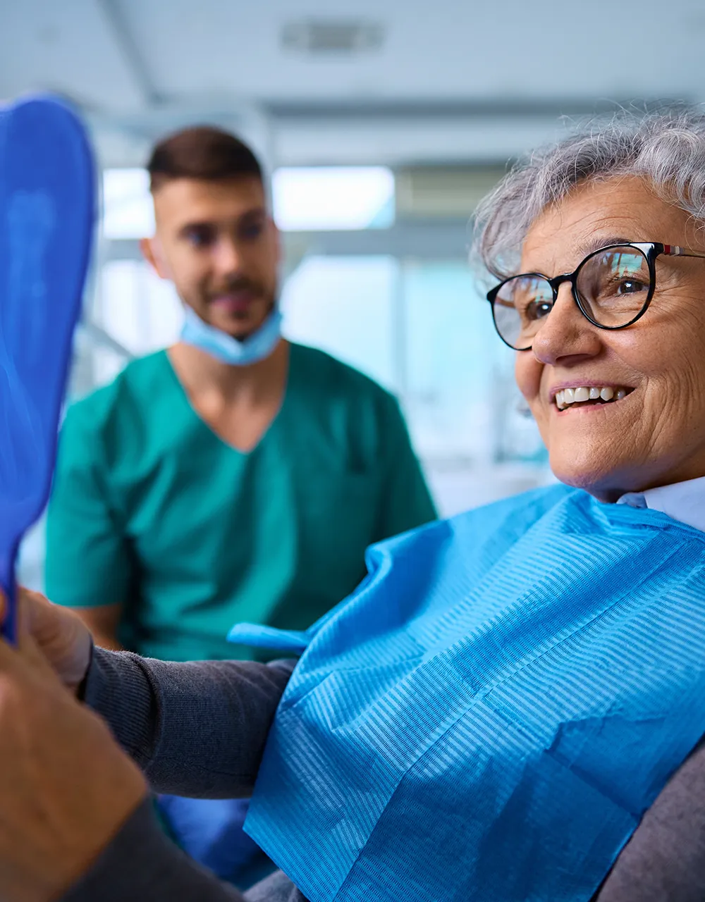 Smiling elderly woman wearing glasses holding a mirror, examining her new dentures, with a dental professional standing in the background.