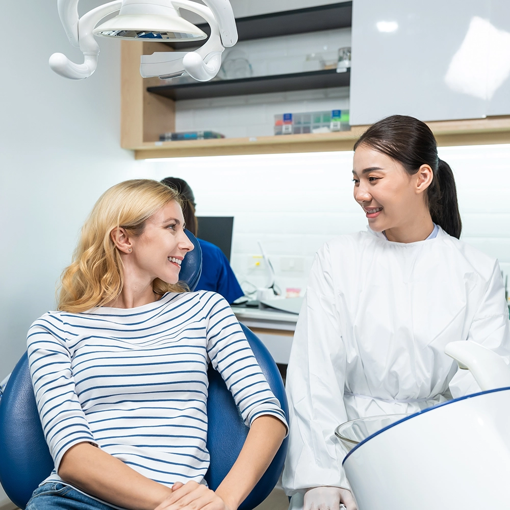 Dentist in a white coat discussing treatment with a smiling blonde patient in a dental chair, showcasing a comfortable and interactive dental visit.